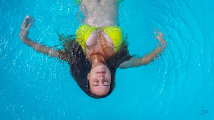 Femme flottant dans une piscine, cheveux longs et mouillés, maillot de bain jaune.